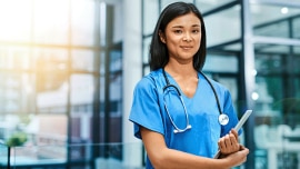 A nurse stand in front of a hospital, smiling at the camera