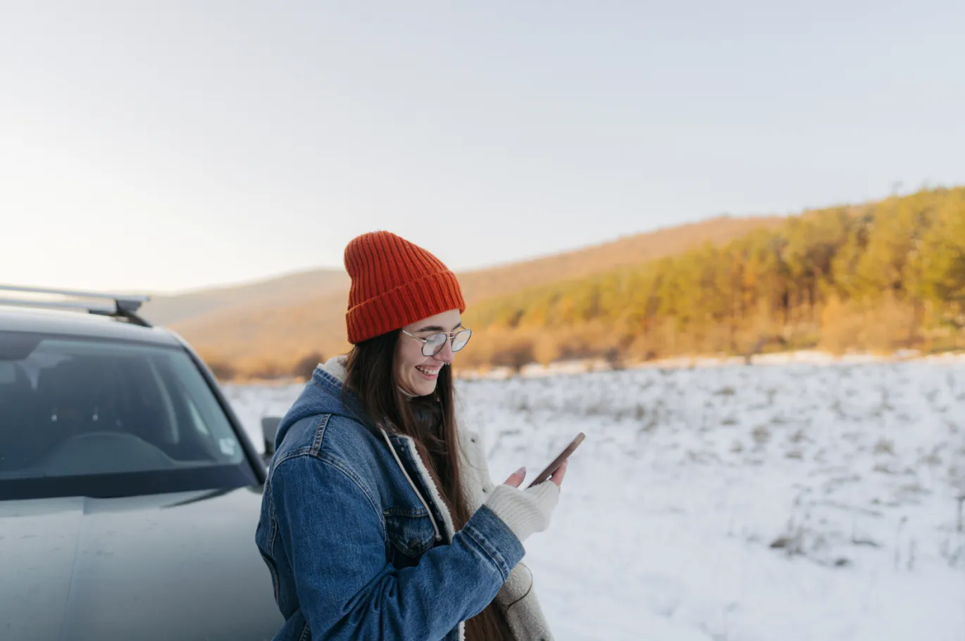 A woman using her phone standing by a car on a snowy street