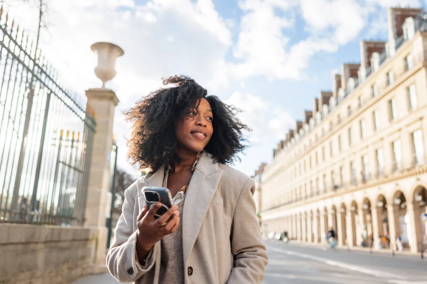 Curly-haired young woman walking through a European city while holding a mobile phone.