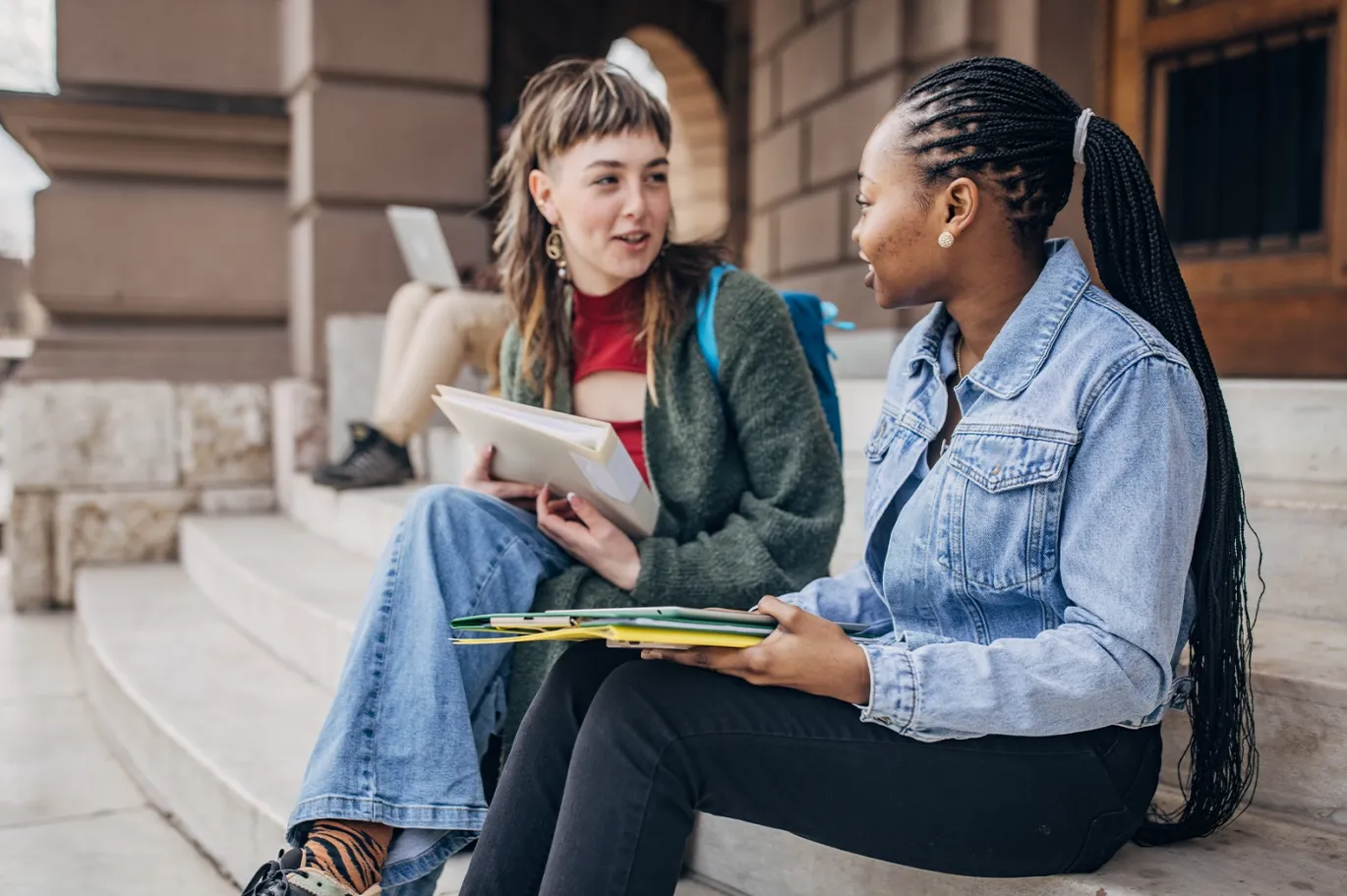 Two students studying together for IELTS exam using notes and a laptop.