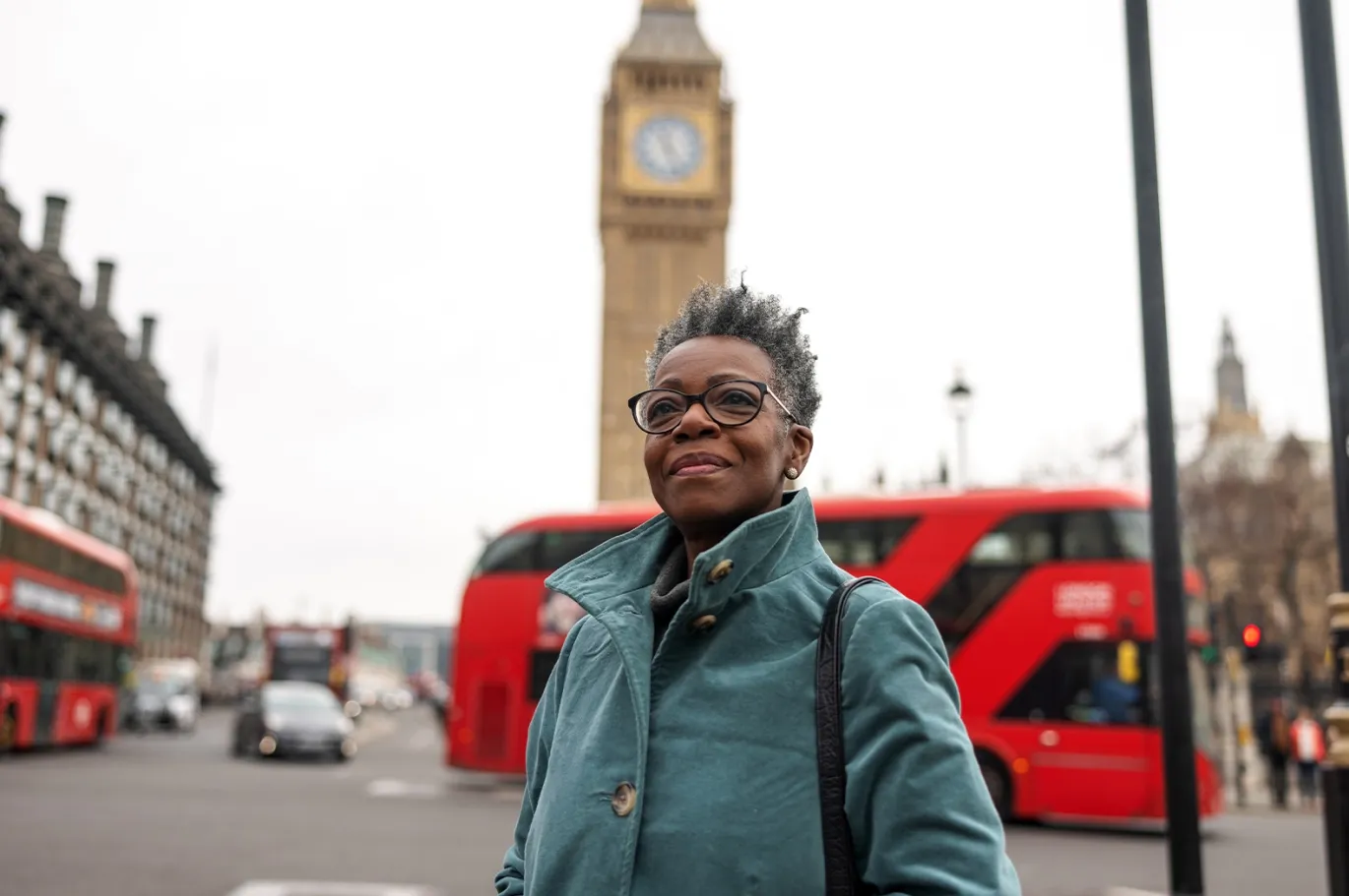 Smiling woman walking near Big Ben in London with red buses in the background.