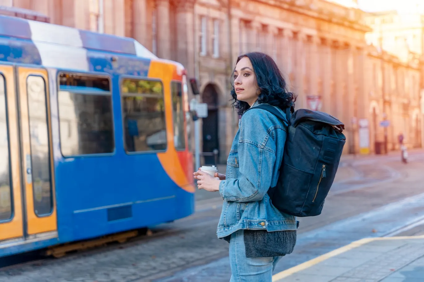 Young woman with a backpack waiting beside a tram in the city going to study abroad.