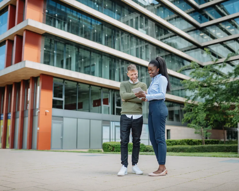 Two students standing and discussing study materials near a campus building