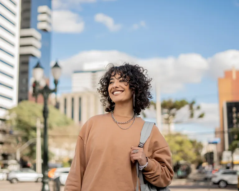 Curly-haired young woman with a backpack smiling while walking through a city street.