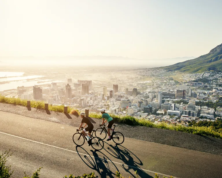 Two cyclists riding along a scenic mountain road overlooking a city.