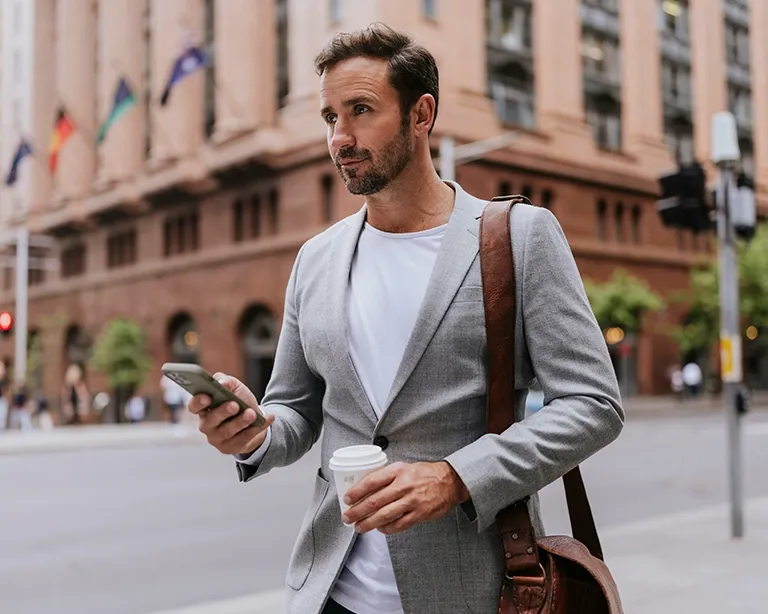 Young man with a shoulder bag holding phone and coffee cup while walking around in the street.