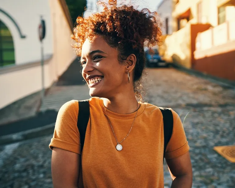 Young woman with a backpack smiling while walking outdoors on a sunny day.