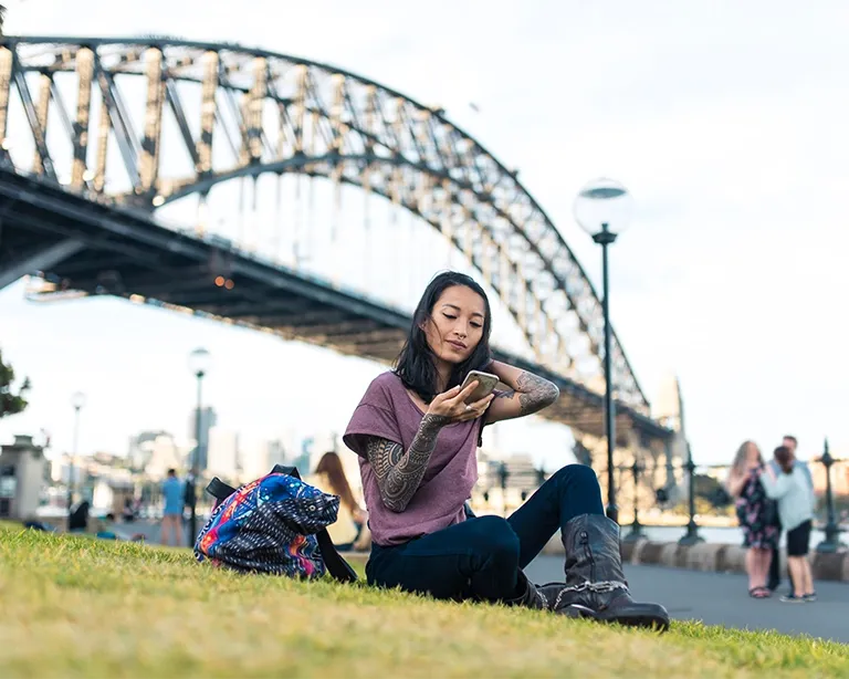 Young woman sitting outdoors near Sydney Harbour Bridge with a backpack.