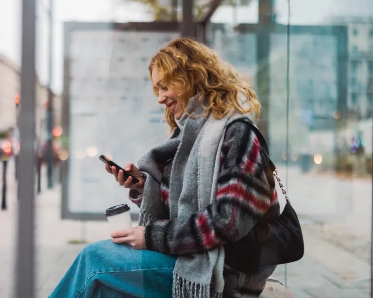 Woman sitting at a bus stop while using her mobile phone and holding a coffee cup.