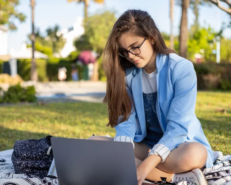 Young woman studying online outdoors using a laptop and IELTS study materials.