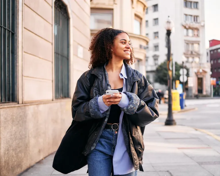 Young woman walking through a city street while holding a mobile phone